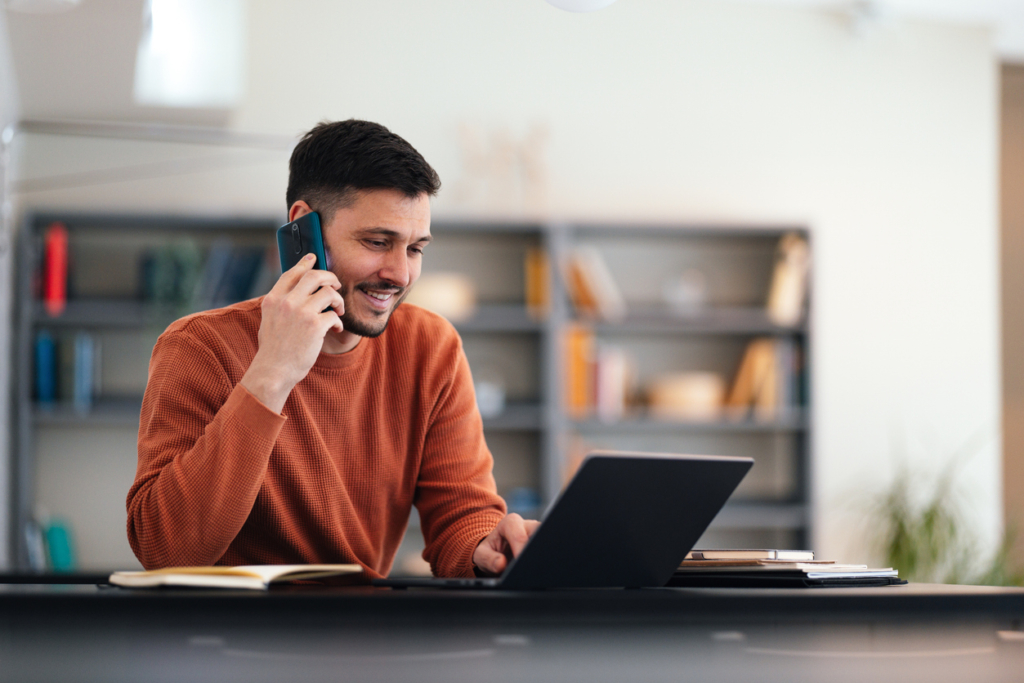 Glücklicher Mann, der mit einem BLV-Mitglied telefoniert, dabei etwas auf der Tastatur seines Laptops tippt und an seinem Büroschreibtisch sitzt.