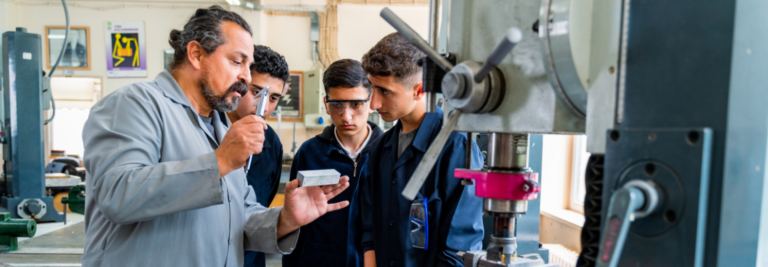 Ein Technsicher Lehrer steht mit drei Schülern in einer Werkstatt und erklärt ihnen anschaulich etwas.