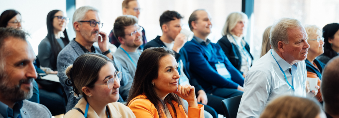 Personen sitzen im Publikum einer Veranstaltung. Die Männer und Frauen hören interessiert zu, einige lächeln. Sie tragen eine Teilnehmerkarte um den Hals.