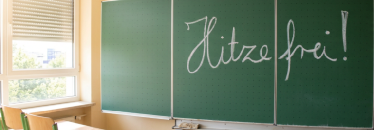 Schultafel mit der Aufschrift: Hitzefrei!", davor Stühle.