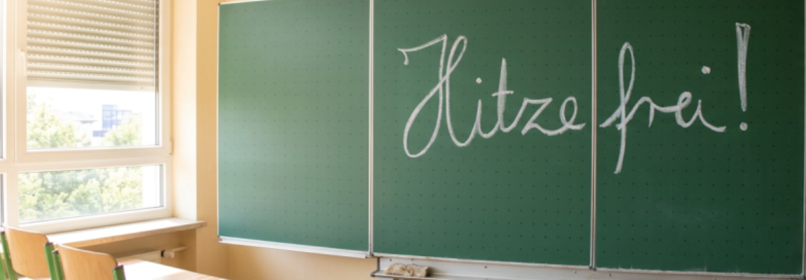 Schultafel mit der Aufschrift: Hitzefrei!", davor Stühle.